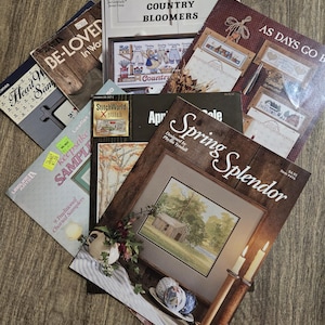 May include: A collection of seven cross-stitch pattern booklets featuring various designs, including Spring Splendor, Country Bloomers, and Heart-Warmed Samplers. The covers display finished embroidery projects, instructional text, and brand logos.