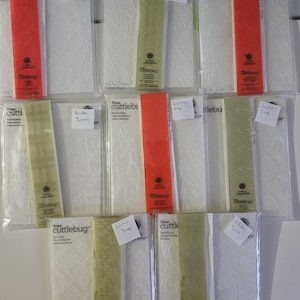 May include: A collection of Cricut Cuttlebug embossing folders in clear plastic packaging. The folders feature various designs and are labeled with names like "Elegant Art" and "Joyful Imp." Each package has a colored strip, including red and green.