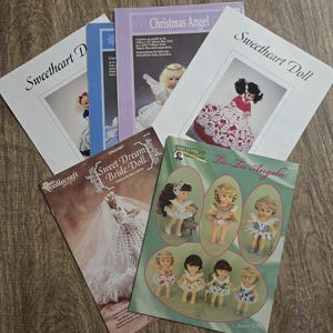 May include: A collection of vintage crochet pattern booklets, including titles like "Sweetheart Doll," "Christmas Angel," and "Sweet Dream Bride Doll." The covers feature illustrations of dolls and angels, with text in black and blue.
