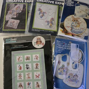 May include: A collection of five craft kits in plastic packaging, including cross-stitch patterns for baby bibs, a crib sheet, a pillowcase, and a stamped quilt featuring various woodland animals like owls, deer, and foxes.