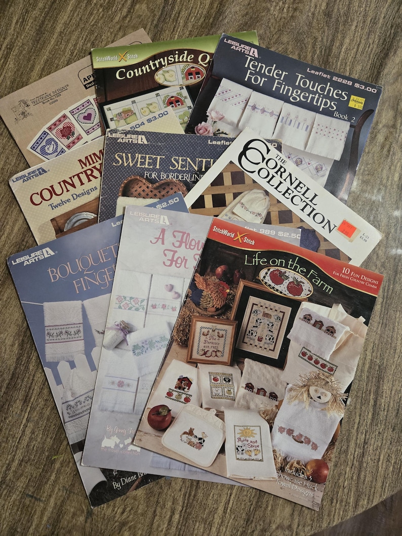 May include: A collection of cross-stitch pattern booklets with titles like "Tender Touches for Fingertips" and "Life on the Farm." The covers feature images of embroidered designs, including floral patterns and country scenes. The booklets are arranged on a wooden surface.