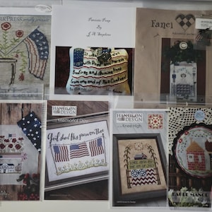 May include: Collection of cross-stitch patterns featuring patriotic themes. Designs include American flags, floral arrangements, and text. The patterns are displayed in clear plastic sleeves, showcasing various styles and designs.