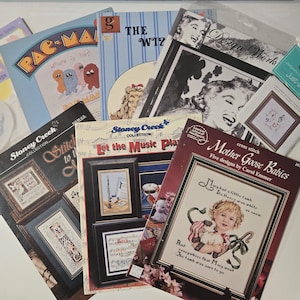 May include: A collection of cross-stitch pattern books, including titles like "Care Bears BABY Sweet Things," "PAC-MAN," and "Mother Goose Babies." The covers feature colorful illustrations and text, with a variety of designs and themes.