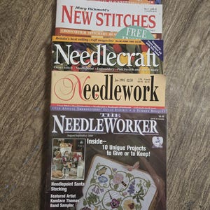 May include: A stack of vintage needlework magazines. Titles include "New Stitches," "Needlecraft," and "Needleworker." The magazines feature colorful covers with text and illustrations related to cross-stitch, embroidery, and needlepoint projects. The magazines are stacked on a wooden surface.