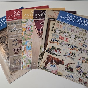 May include: A collection of six Sampler & Antique Needlework magazines arranged in a fan shape. Each magazine cover features colorful cross-stitch embroidery designs, including alphabets, floral wreaths, animals, and historical patterns with text.