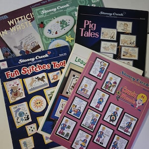 May include: A collection of Stoney Creek cross-stitch pattern books, featuring titles like "Fun Stitches Too!" and "Pig Tales." The covers display colorful cross-stitch designs of animals, characters, and quotes. The books are arranged on a white surface.