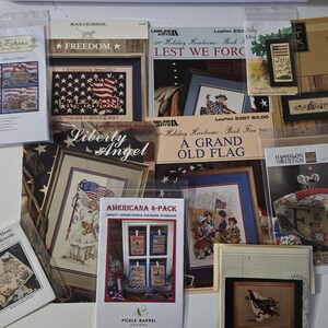 May include: A collection of cross-stitch patterns and leaflets with patriotic themes. Designs include American flags, historical figures, and Americana motifs. The color palette features red, white, and blue.