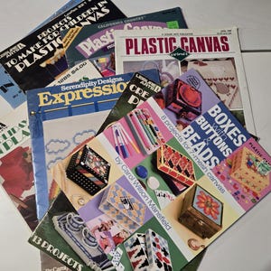May include: A collection of stacked Leisure Arts craft booklets featuring plastic canvas projects. The top booklet is titled Boxes with Buttons & Beads by Carol Wilson Mansfield, displaying various decorated boxes, beads, and buttons on its cover.