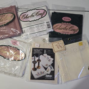May include: Assortment of Charles Craft bread covers and table linens in various colors, including pink, black, and cream. Some items are labeled with "Country Cottage Table Linens" and "Crafts Pride". The linens are packaged in clear plastic.