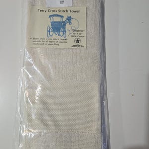 May include: A cream-colored Terry Cross Stitch Towel, packaged in clear plastic. The towel features a three-inch cross-stitch border and is labeled "Empress", measuring 16 x 26 inches. The label also includes a blue illustration of a horse-drawn carriage.
