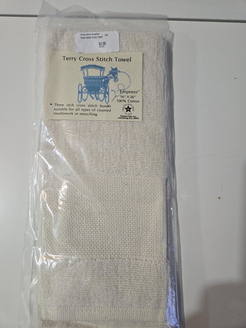 May include: A cream-colored Terry Cross Stitch Towel, packaged in clear plastic. The towel features a three-inch cross-stitch border and is labeled "Empress", measuring 16 x 26 inches. The label also includes a blue illustration of a horse-drawn carriage.