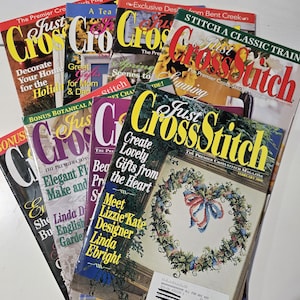 May include: A collection of Just CrossStitch magazines stacked together. The covers feature various cross-stitch patterns, including a floral heart wreath, and text such as 'Create Lovely Gifts from the Heart', 'Meet Designer Linda Ebright', and 'Stitch a Classic Train'.