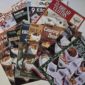 May include: A collection of vintage craft booklets featuring designs for bread cloths and kitchen linens. The covers display images of embroidered cloths, baked goods, and holiday themes. Titles include "25 Bread Cloths" and "Christmas Towels & Bread Cloths."