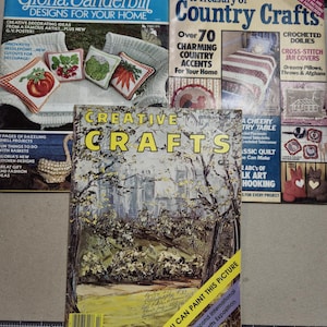 May include: Three vintage craft magazines with colorful covers. The top magazine is titled "Gloria Vanderbilt Designs for Your Home" and features a crocheted white hammock with colorful pillows. The middle magazine is titled "A Treasury of Country Crafts" and features a crocheted white and red blanket. The bottom magazine is titled "Creative Crafts" and features a painting of a country road with trees and a yellow field.