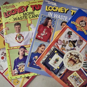 May include: A collection of four Looney Tunes Leisure Arts cross stitch leaflets featuring various character designs, including Marvin the Martian, Bugs Bunny, and the Tasmanian Devil, alongside a round clock face with a Tasmanian Devil graphic.