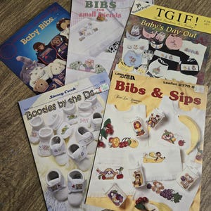 May include: A collection of vintage baby-themed cross-stitch pattern booklets. The covers feature images of baby bibs, booties, and other baby-related items. The booklets have titles like "Baby Bibs," "Bibs for Small Friends," and "Bibs & Sips."