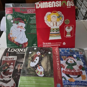 May include: A collection of Christmas felt applique kits, including a Santa Claus stocking, a tree top angel, a snowman stocking, and a penguin wall hanging. The kits feature various colors and designs, perfect for holiday crafting.