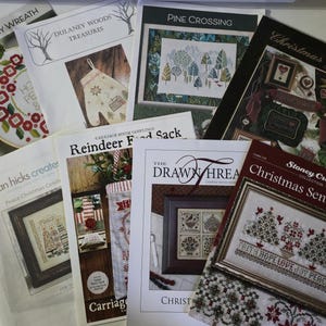 May include: A collection of seven cross-stitch pattern booklets featuring various holiday and seasonal designs. Titles include Peaceful Posy Wreath, Dulaney Woods Treasures, Pine Crossing, Christmas Jewels, Peace Christmas Candle, Reindeer Food Sack, and Christmas Sentiments.