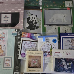 May include: A collection of cross-stitch kits, featuring various designs. Kits include images of a teddy bear, pandas, polar bears, a baby with balloons, and a wine-themed design. The kits are packaged in plastic.