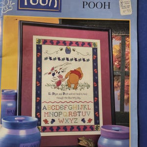 May include: Cross stitch pattern featuring Winnie the Pooh and Piglet walking through a forest with a basket of honey. The pattern includes the alphabet and the text "So Piglet and Pooh walked hand in hand through the blustery day."
