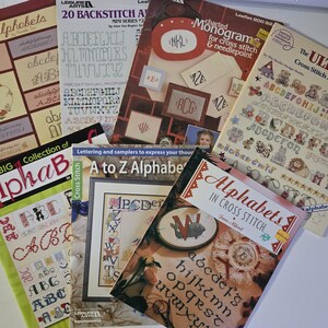 May include: A collection of seven instructional books for cross-stitch and needlepoint, featuring various alphabet designs, monograms, and lettering patterns. The covers display titles such as Script Alphabets, 20 Backstitch Alphabets, Charted Monograms, The Ultimate Cross Stitch Alphabet Book, AlphaB, A to Z Alphabets, and Alphabets in Cross Stitch.