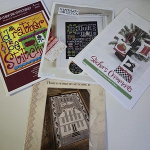 May include: A collection of cross-stitch pattern booklets. The booklets feature various designs, including the text "I'd Rather Be Stitching", a framed house, and Christmas ornaments. The color palette includes burgundy, white, and green.