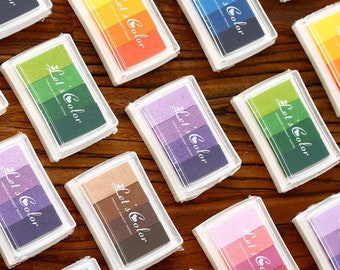 Puede incluir: Una colección de almohadillas de tinta rectangulares en estuches de plástico transparente. Cada almohadilla presenta tres tonos de color, con el texto "Let's Color" y "Premium Pigment Pad". Las combinaciones de colores incluyen azul, verde, morado y rosa.