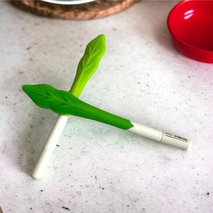 May include: A white marker with green leaf-shaped covers on each end. The marker is lying on a white surface.