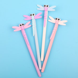 May include: Four colorful pens with dragonfly toppers. The dragonflies are pink, blue, white, and purple. The pens are all black.