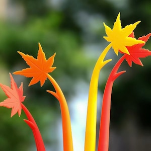 May include: Four orange and yellow maple leaf shaped paperweights with long stems. The stems are arranged in a fan shape.
