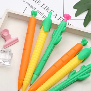 May include: A collection of novelty pens shaped like vegetables. The pens include orange carrots, yellow corn, and green cacti. The pens are arranged on a white tray with the text "LIKE A F" visible in the background.
