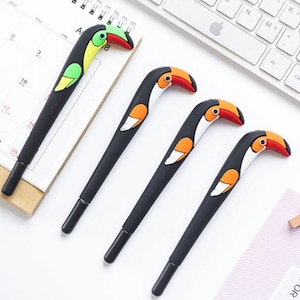 Puede incluir: Cuatro bolígrafos con forma de tucán con cuerpos negros y cabezas de tucán de colores. Los tucanes tienen picos naranja, amarillo y verde y ojos negros.
