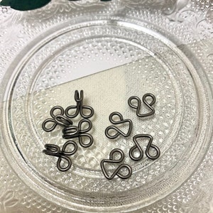 May include: A close-up of a group of silver metal hook and eye closures on a white surface. The closures are arranged in a random pattern.