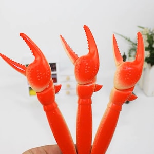 May include: Three red plastic crab claw shaped pens. The claws are open and the pens are ready to write.