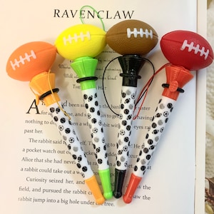 Puede incluir: Cuatro bolígrafos de colores con forma de balón de fútbol con lunares blancos y negros. Los bolígrafos tienen un balón de fútbol verde, naranja, negro y rojo en la parte superior. El texto "RAVENCLAW" es visible en el fondo.