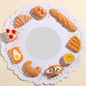 Può includere: Un set di nove ciondoli per alimenti in miniatura, tra cui un croissant, una fetta di pizza, un waffle, una baguette, un dolce, un croissant, una fetta di pane con un uovo, un dolce con un vortice di crema e una piccola ciotola con un ripieno giallo. I ciondoli sono disposti su un pizzo bianco.