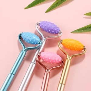 Puede incluir: Cuatro rodillos faciales con mangos y cabezales de rodillo de diferentes colores. Los cabezales de rodillo son morado, azul, rosa y naranja. Los mangos son plateados, oro rosa y azul.