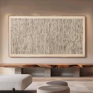 May include: A living room interior with a white couch, two round ottomans, and a long wooden shelf with a black and white abstract art piece hanging on the wall above it. The walls are a light beige color and the floor is a light brown wood.