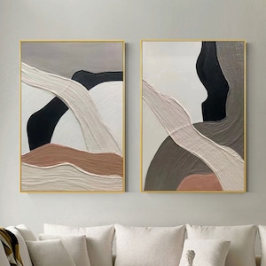 Puede incluir: Dos piezas de arte mural rectangulares con composiciones abstractas de pinceladas gruesas y texturizadas en tonos negro, blanco, beige y marrón apagado. Cada lienzo está enmarcado con un borde dorado fino, mostrando formas orgánicas superpuestas.