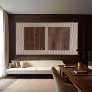 Puede incluir: Gran obra de arte mural abstracto con dos cuadrados marrones texturizados sobre un fondo crema con l&iacute;neas verticales. La obra est&aacute; enmarcada en dorado y cuelga sobre un sof&aacute; blanco. La habitaci&oacute;n tiene paneles de madera oscura.