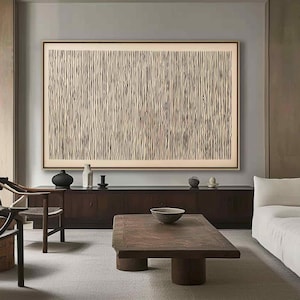 May include: Large framed abstract artwork with a beige background and vertical dark brown lines. The artwork is displayed above a dark wood cabinet with decorative items. A wooden coffee table and a white sofa are in the foreground.