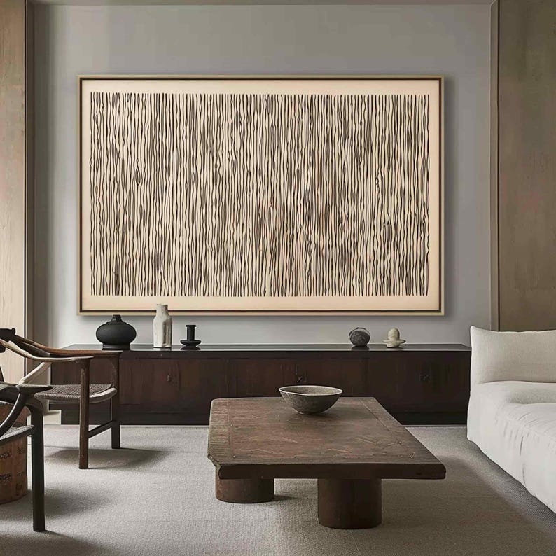 May include: Large framed abstract artwork with a beige background and vertical dark brown lines. The artwork is displayed above a dark wood cabinet with decorative items. A wooden coffee table and a white sofa are in the foreground.