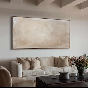 May include: A large, rectangular abstract painting with a brown frame. The artwork features a textured surface with a blend of beige and off-white hues. The painting hangs above a white sofa with beige pillows.