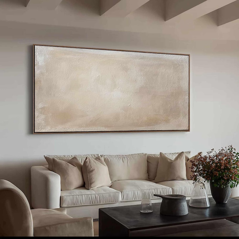 May include: A large, rectangular abstract painting with a brown frame. The artwork features a textured surface with a blend of beige and off-white hues. The painting hangs above a white sofa with beige pillows.