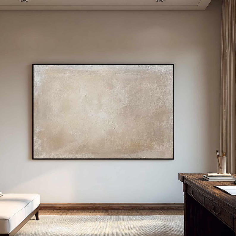 May include: Large abstract painting in a black frame. The artwork features a textured, neutral color palette with shades of beige and cream. The painting hangs on a white wall in a room with a wooden desk and a white sofa.