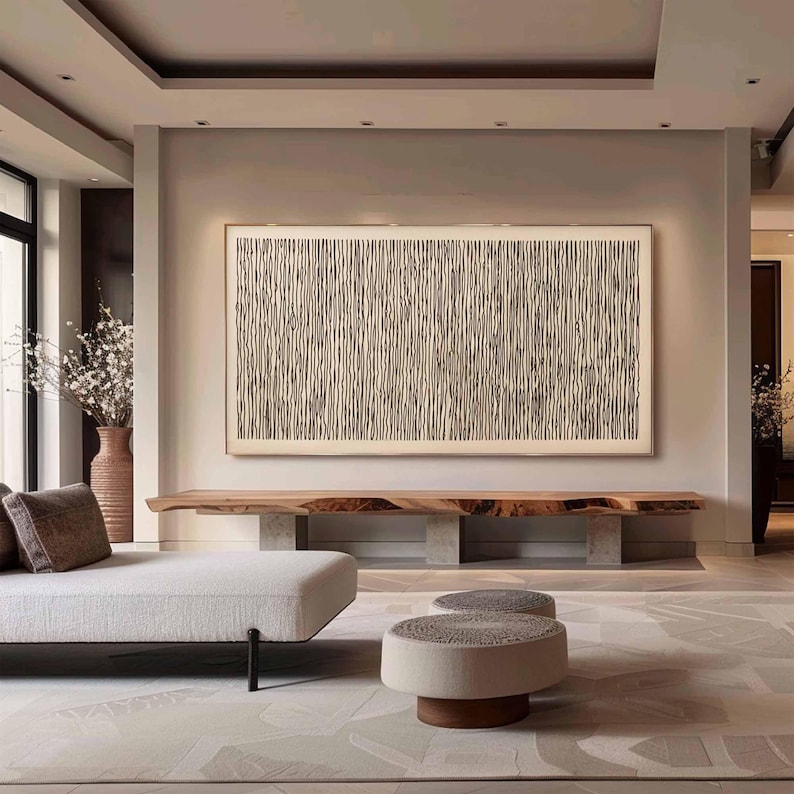 May include: A living room interior with a white couch, two round ottomans, and a long wooden shelf with a black and white abstract art piece hanging on the wall above it. The walls are a light beige color and the floor is a light brown wood.