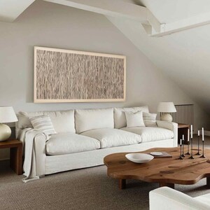 May include: A living room with a large white sofa, a wooden coffee table, and a framed artwork on the wall. The artwork features a beige and brown vertical line pattern. Two white lamps sit on wooden side tables. The room has a neutral color palette.