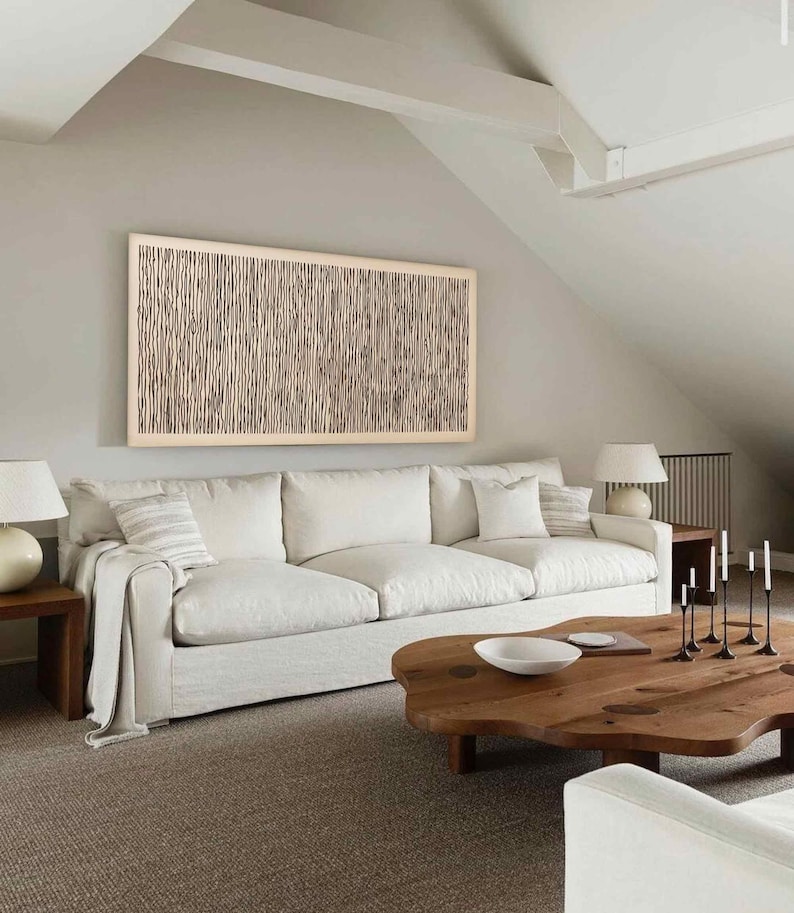May include: A living room with a large white sofa, a wooden coffee table, and a framed artwork on the wall. The artwork features a beige and brown vertical line pattern. Two white lamps sit on wooden side tables. The room has a neutral color palette.