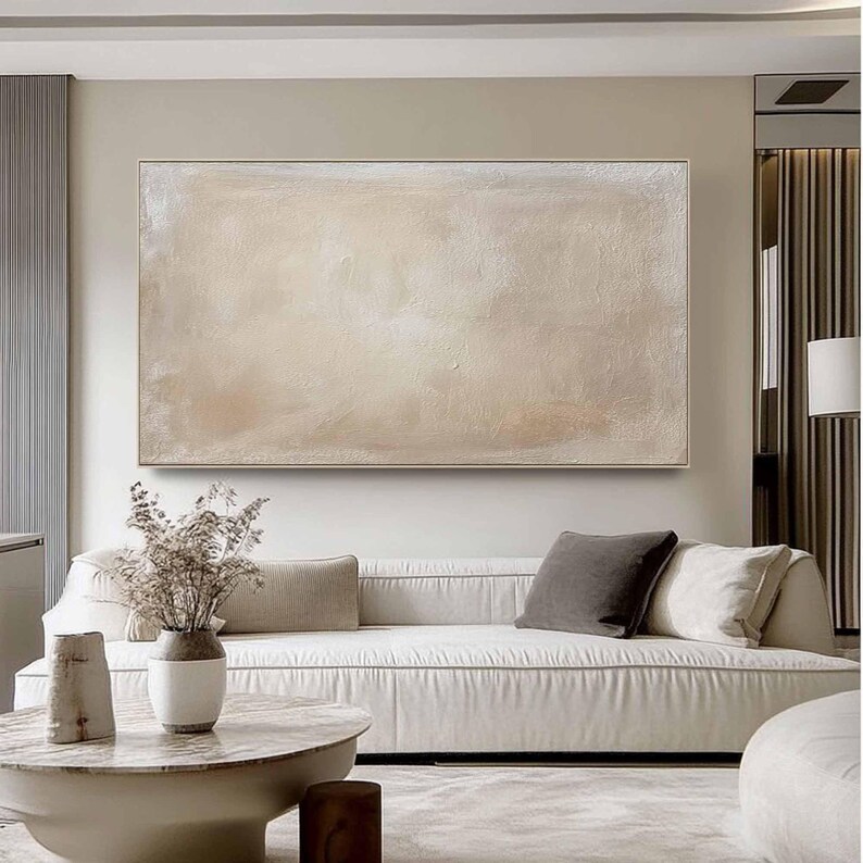 May include: A large, rectangular abstract painting in neutral tones hangs above a white sofa. The artwork features a textured surface with shades of beige, cream, and light brown. The living room setting includes a round coffee table and decorative vase.