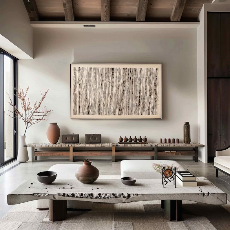 May include: A modern living room with neutral tones. A large, framed artwork with a vertical line pattern hangs above a wooden bench. Decorative vases, bowls, and small sculptures are arranged on the bench and a large, live-edge coffee table. The room has a minimalist aesthetic.